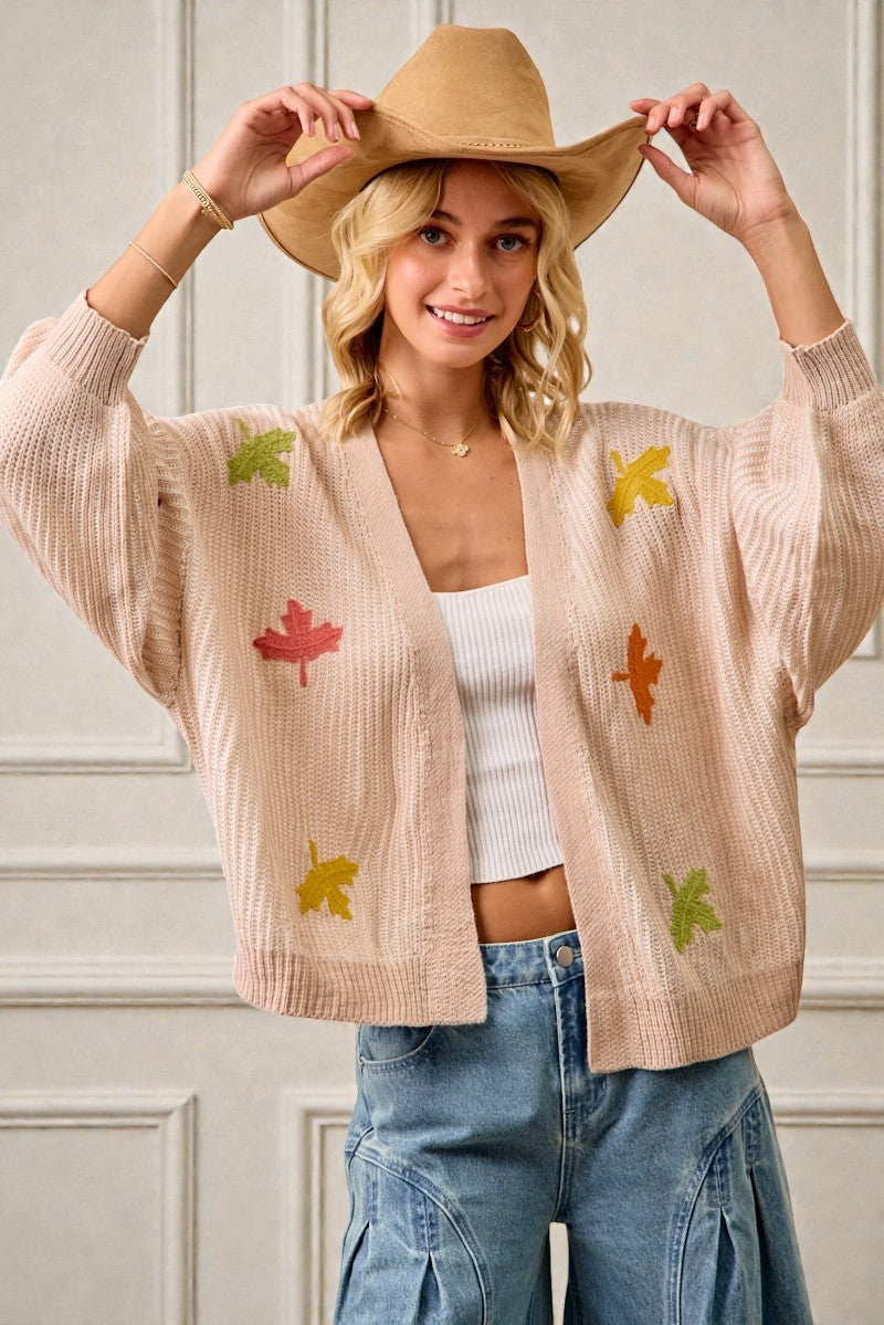 Woman wearing a beige cardigan with leaf patterns, white top, and blue jeans against a neutral background.
