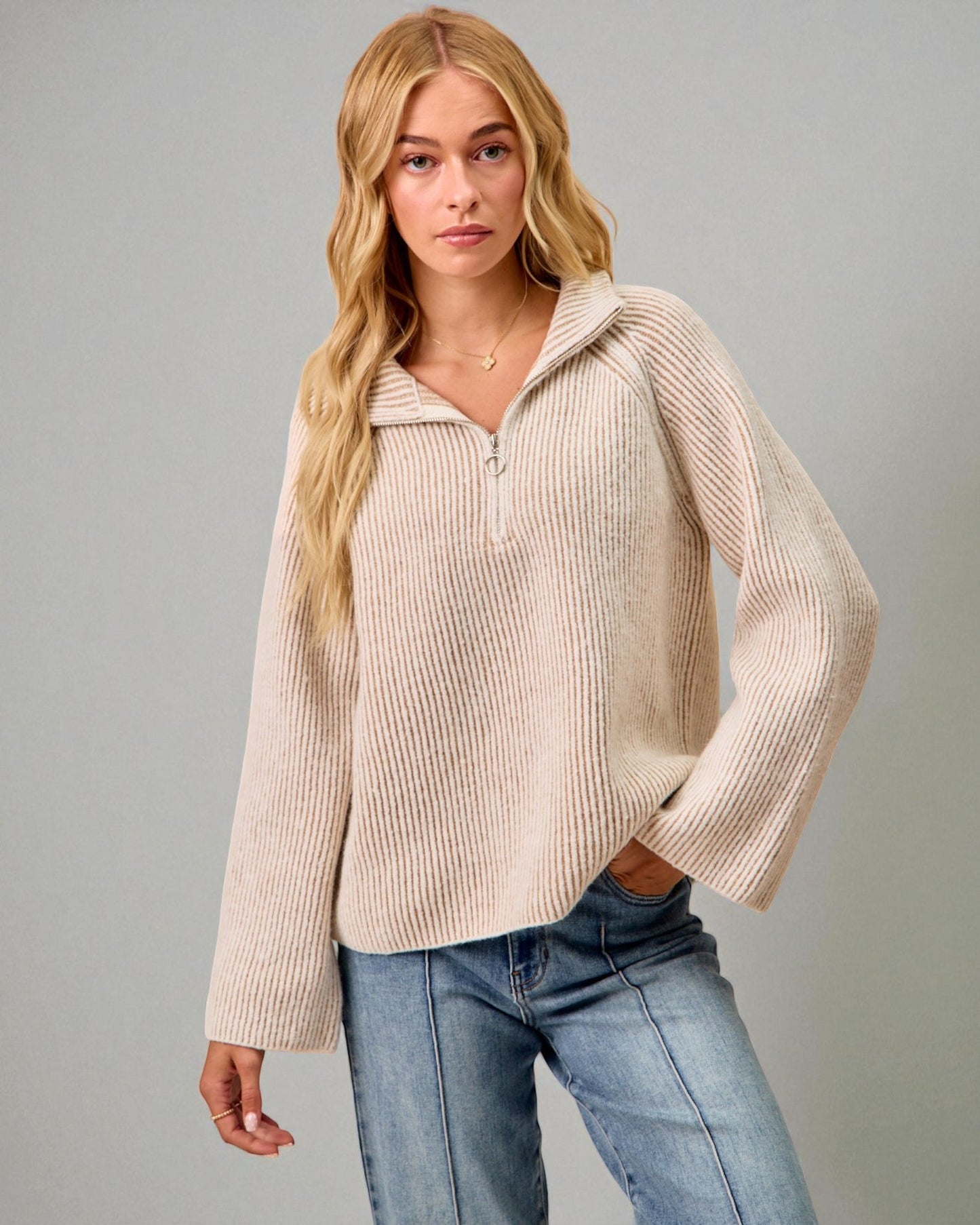 Woman wearing a beige ribbed sweater and blue jeans against a gray background