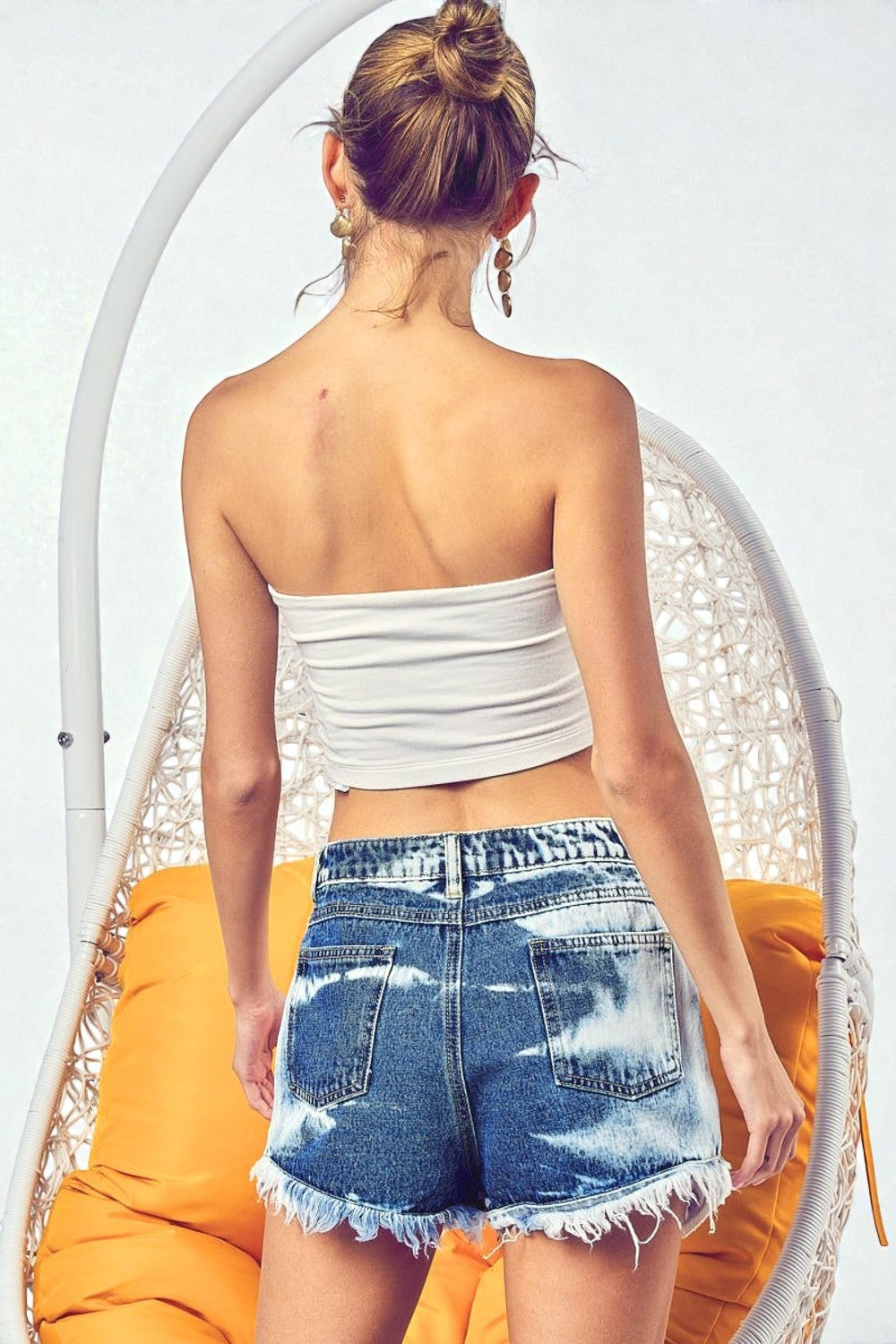 Woman wearing a white strapless top and denim shorts sitting on a white chair with an orange cushion.