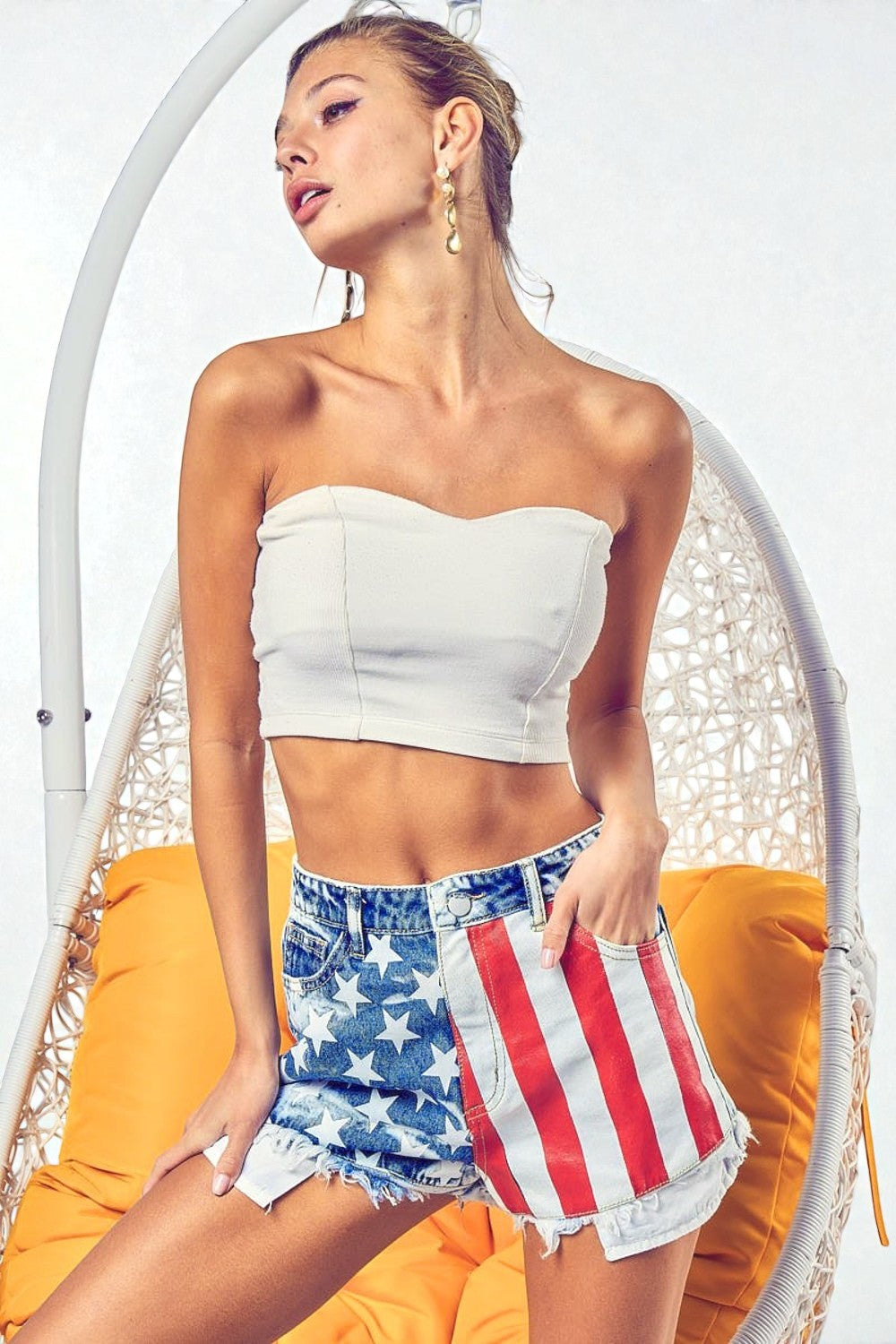 Woman wearing a white strapless top and American flag shorts sitting on a yellow cushioned chair.
