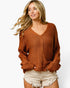 A woman wearing a chunky knit brown V-neck sweater and light beige frayed denim shorts, photographed against a clean white studio background.