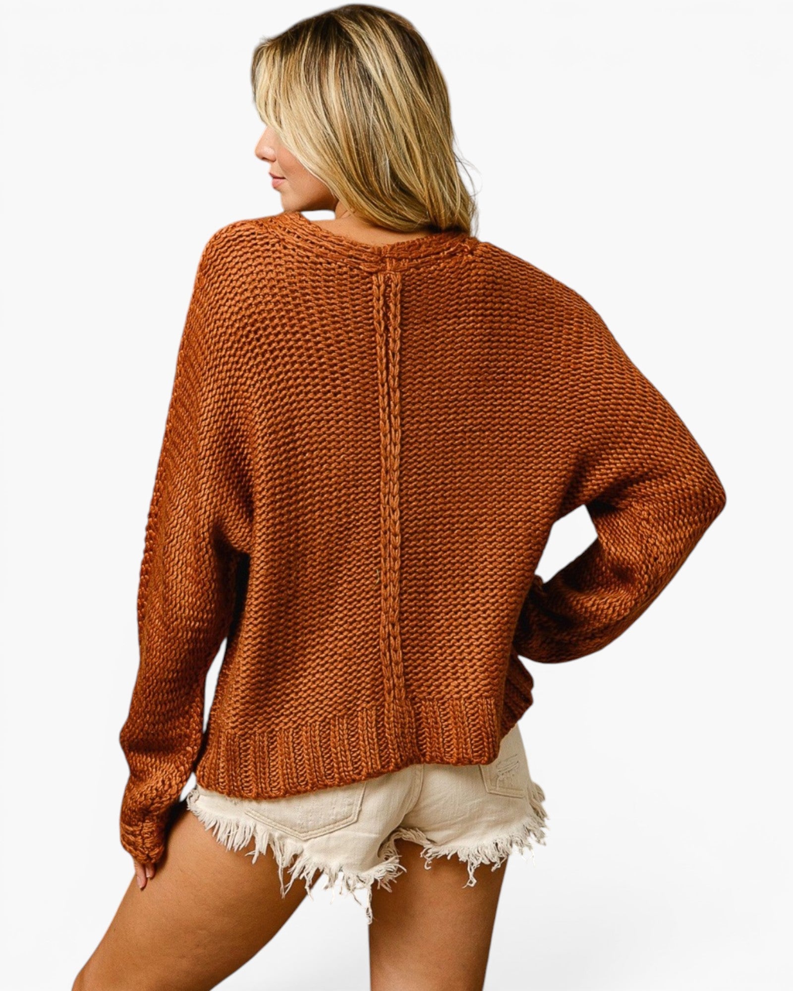 A woman shown from the back wearing a chunky knit crewneck sweater in a warm rust orange color, paired with cream frayed denim shorts, against a plain white background.