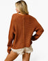 A woman shown from the back wearing a chunky knit crewneck sweater in a warm rust orange color, paired with cream frayed denim shorts, against a plain white background.