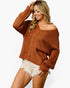 A woman modeling a chunky knit off-the-shoulder sweater in a warm rust brown color, paired with light beige frayed denim shorts, against a plain white background.