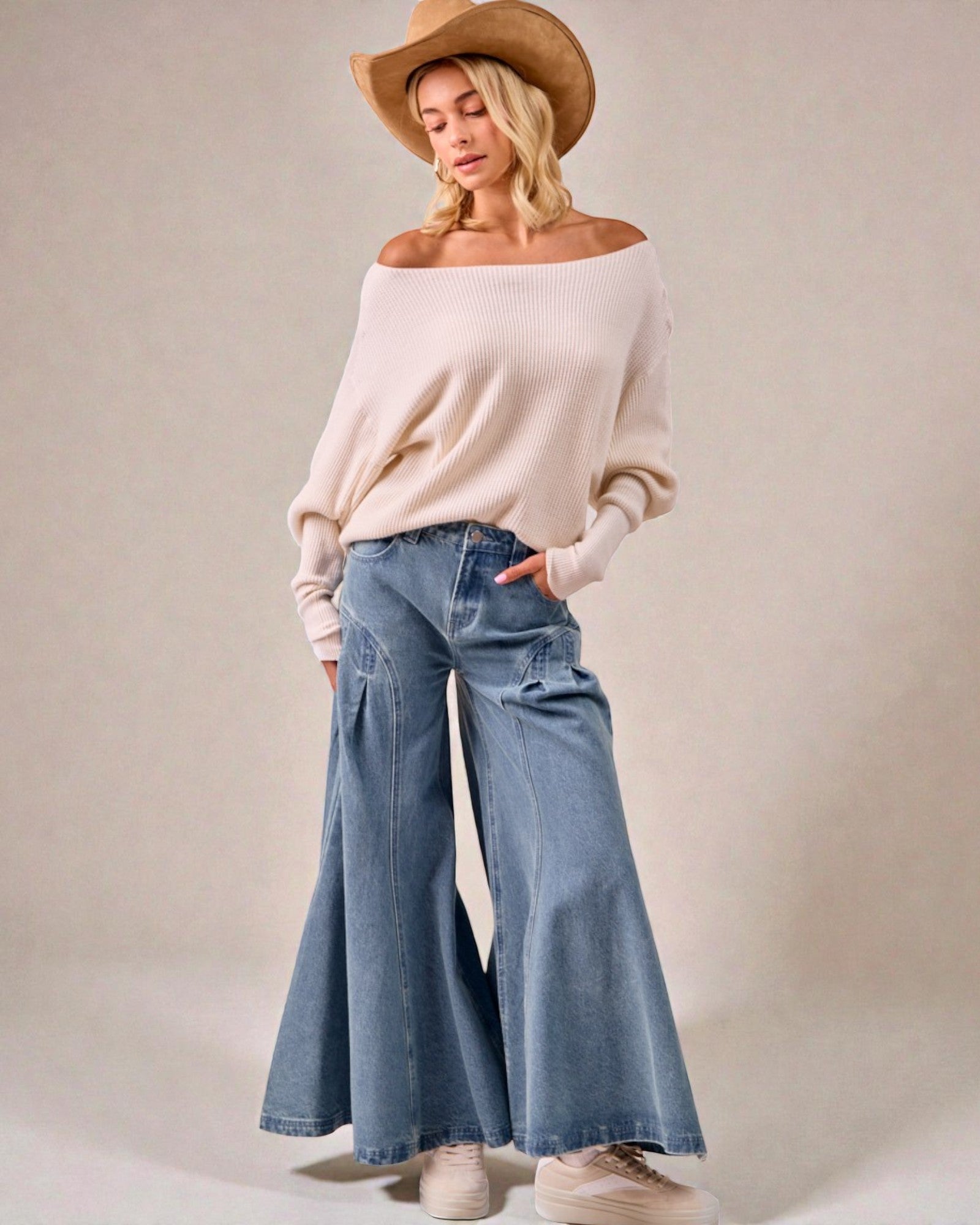 Woman wearing a beige off-shoulder sweater and blue jeans with a beige hat on a plain background