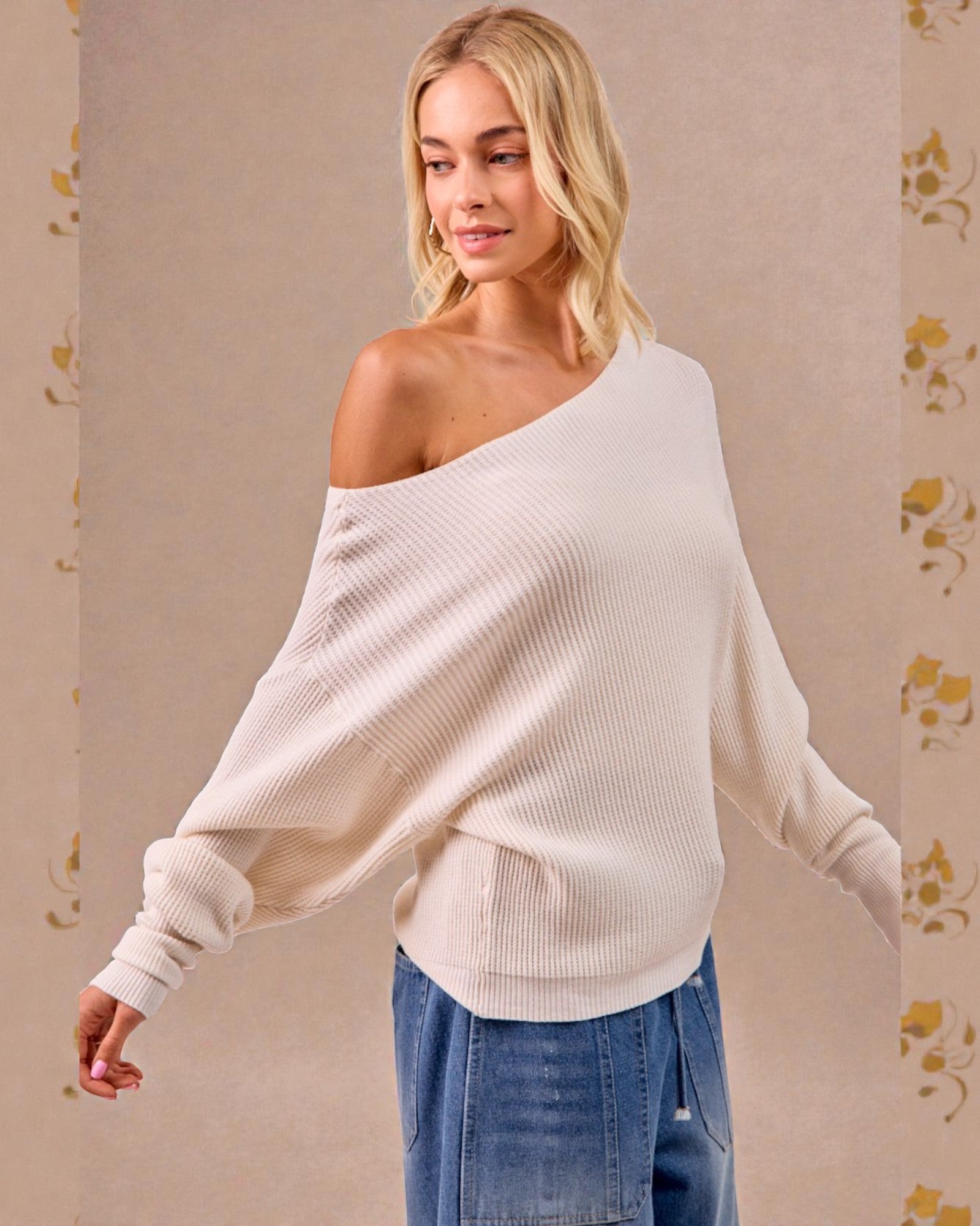 Woman wearing a white off-shoulder sweater and blue jeans against a beige background