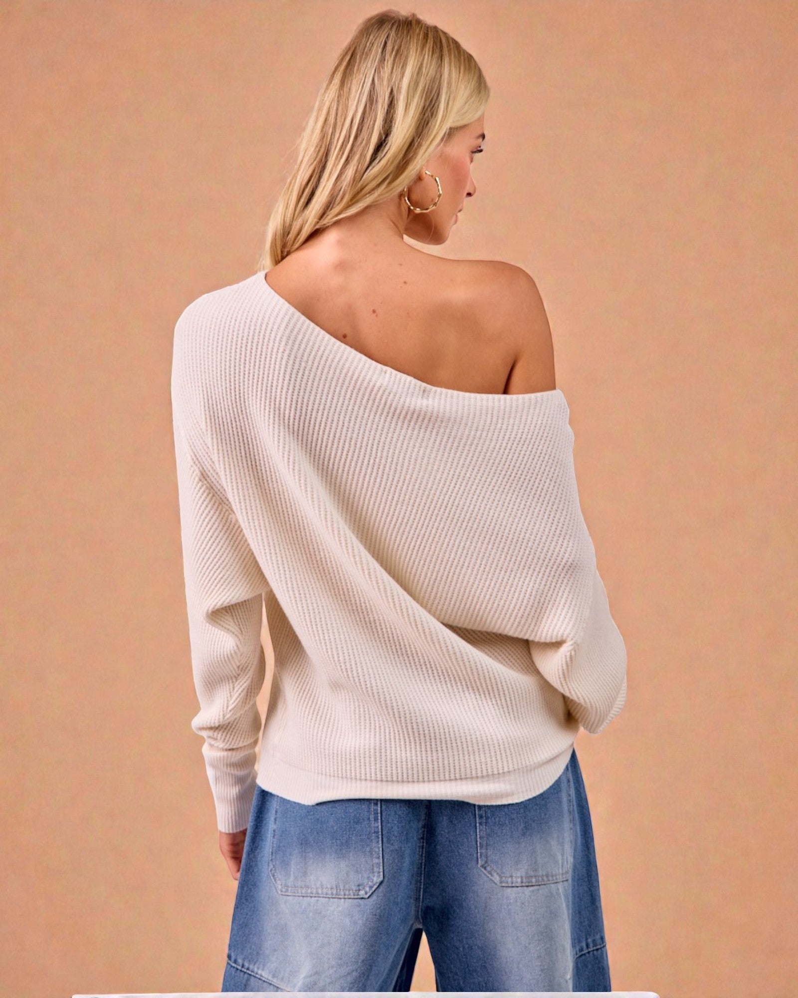Woman wearing a beige off-shoulder sweater and blue jeans against a beige background