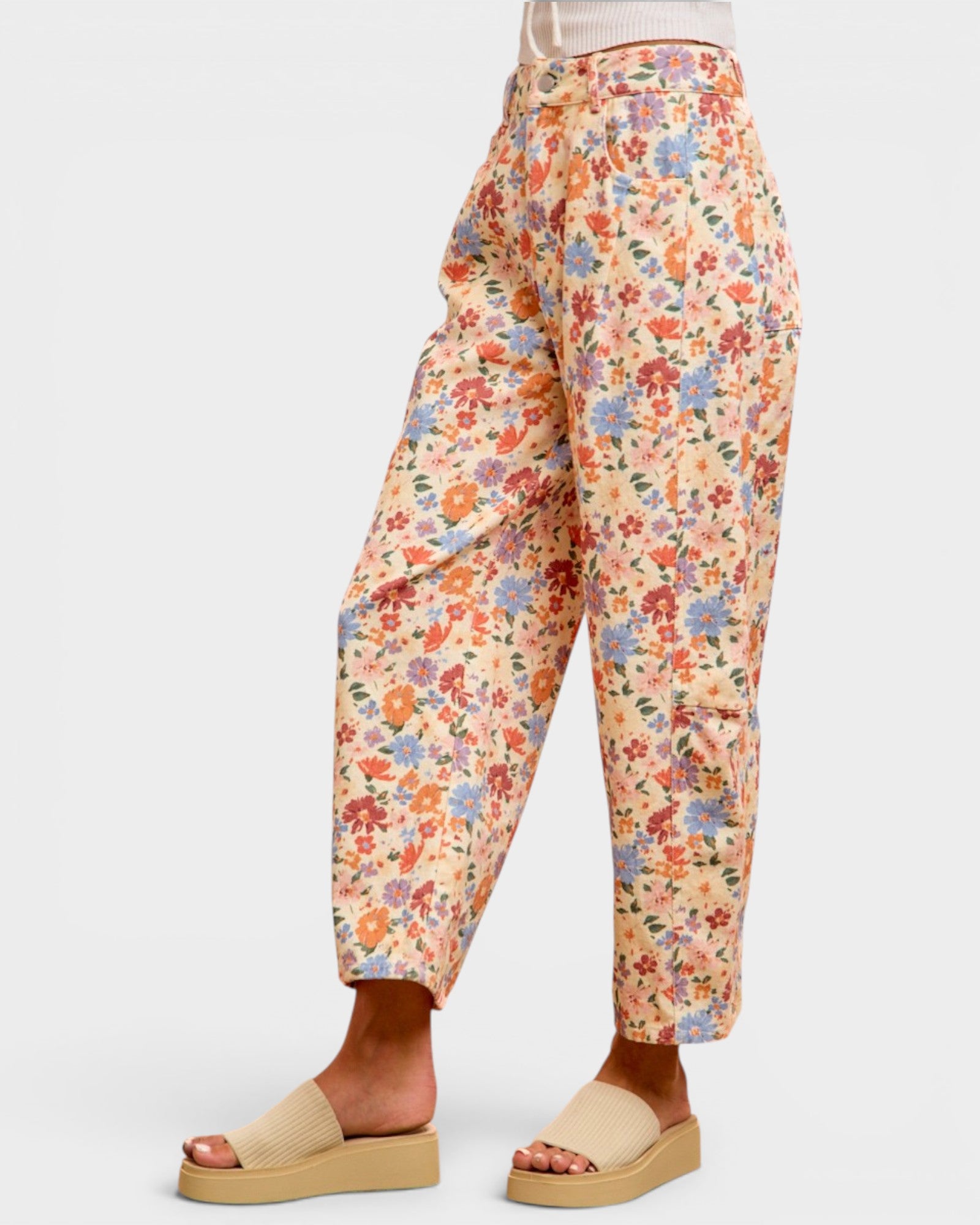 A person wearing floral cropped pants with a beige top and beige slide sandals, shown from the waist down against a light gray studio background.