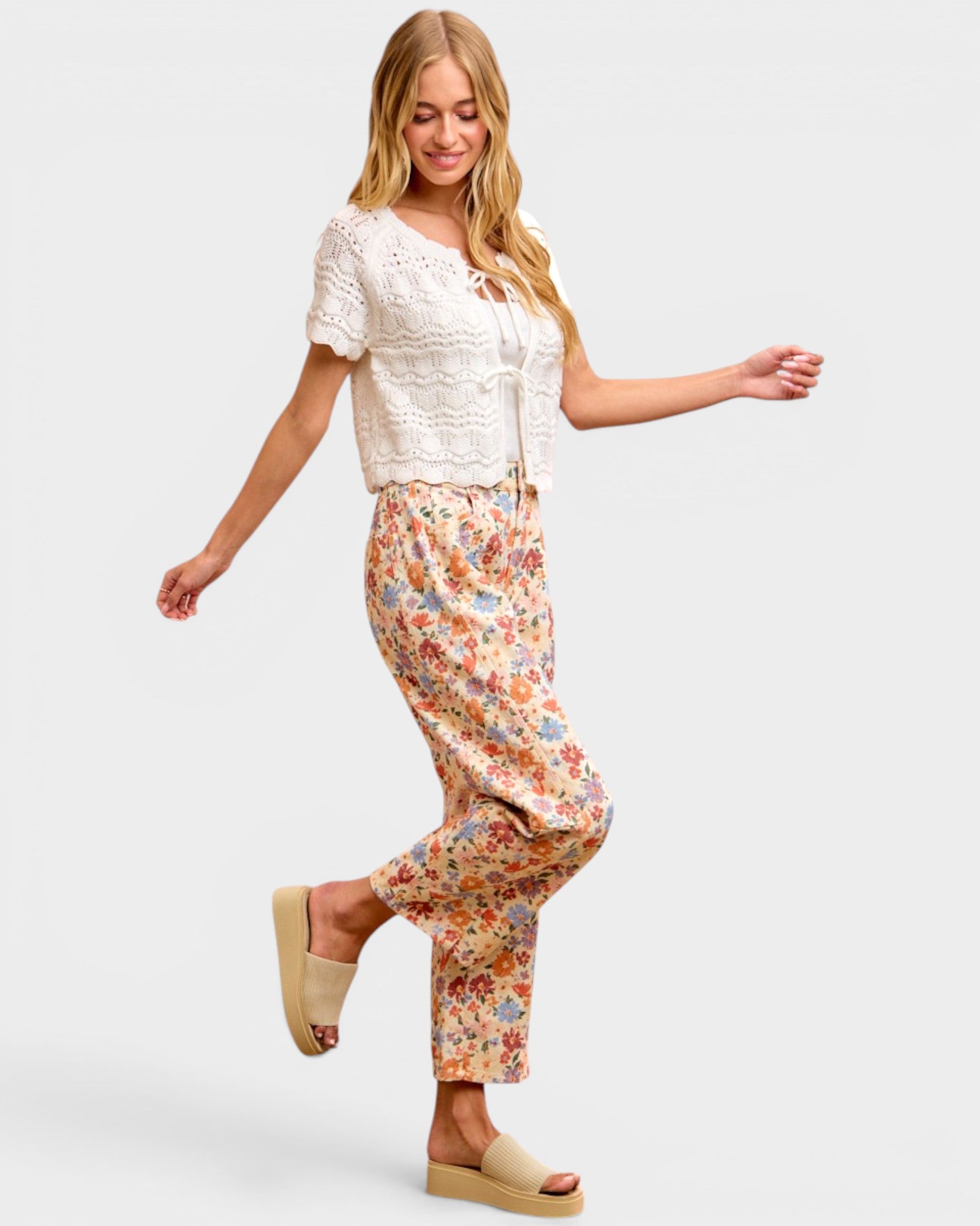 A woman modeling a two-piece outfit consisting of a white short-sleeve crochet knit top and beige floral print pants, paired with beige platform slide sandals, against a light gray studio background.