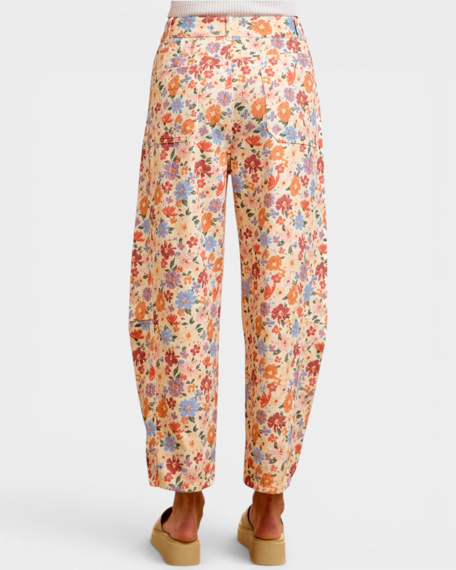 A pair of wide-leg cropped pants with an all-over colorful floral print on a light cream background, shown from the back on a model wearing tan platform sandals against a light gray studio background.