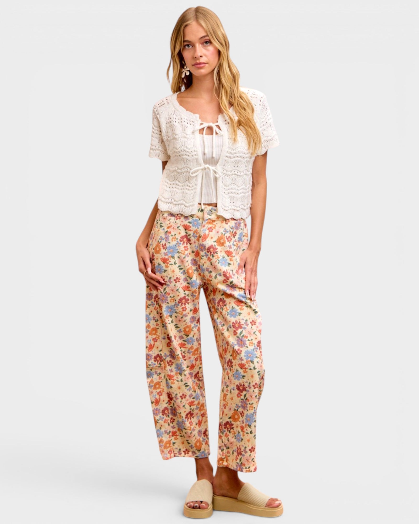 A woman modeling a two-piece outfit consisting of a white short-sleeve lace-trim crop top and beige wide-leg pants with a colorful floral print, paired with beige slide sandals against a light gray background.
