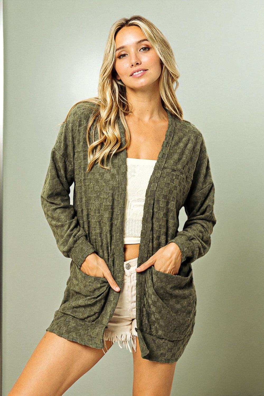 Woman wearing a green cardigan over a white top and shorts on a plain background