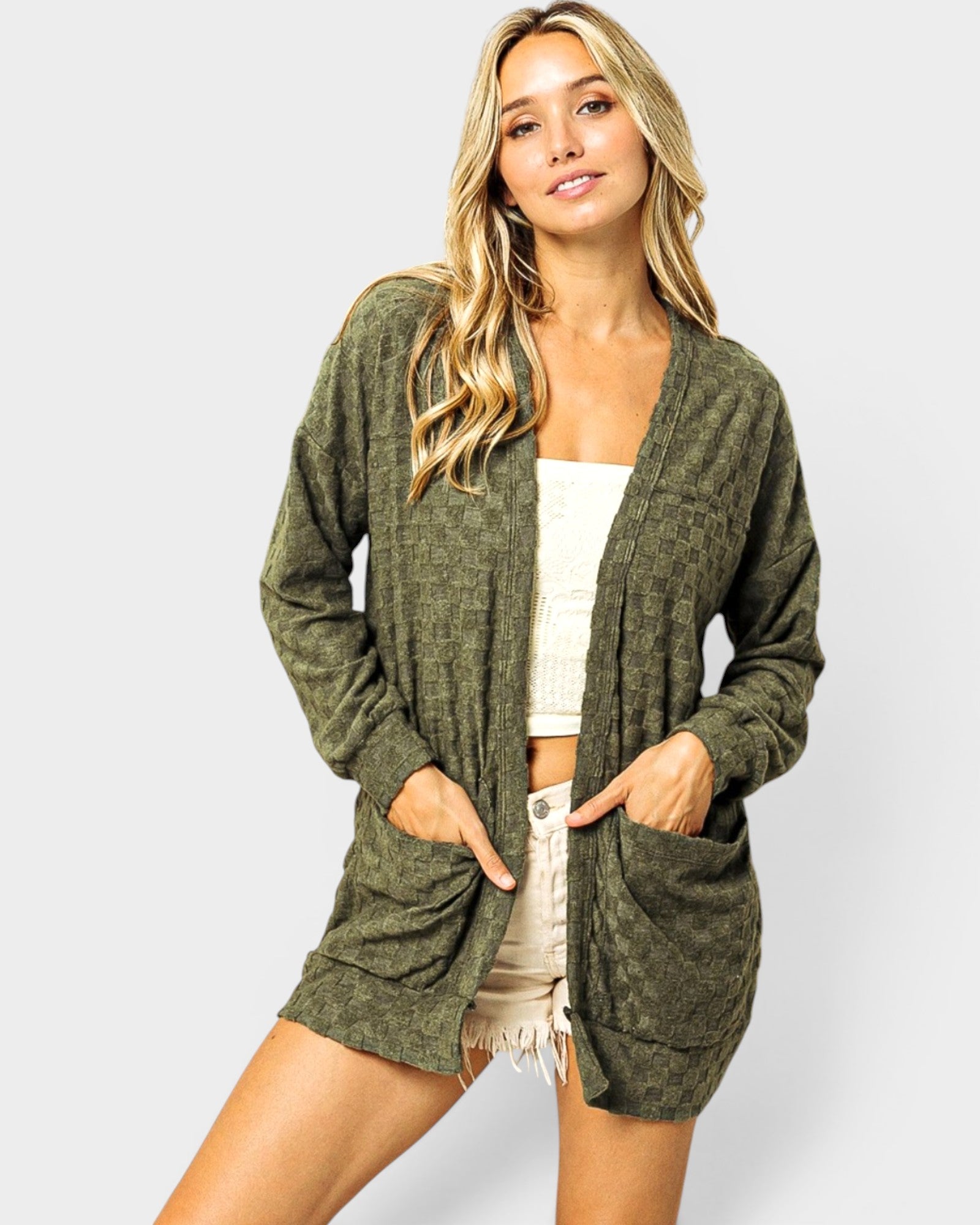 Woman wearing a green cardigan over a white top and beige shorts on a light gray background