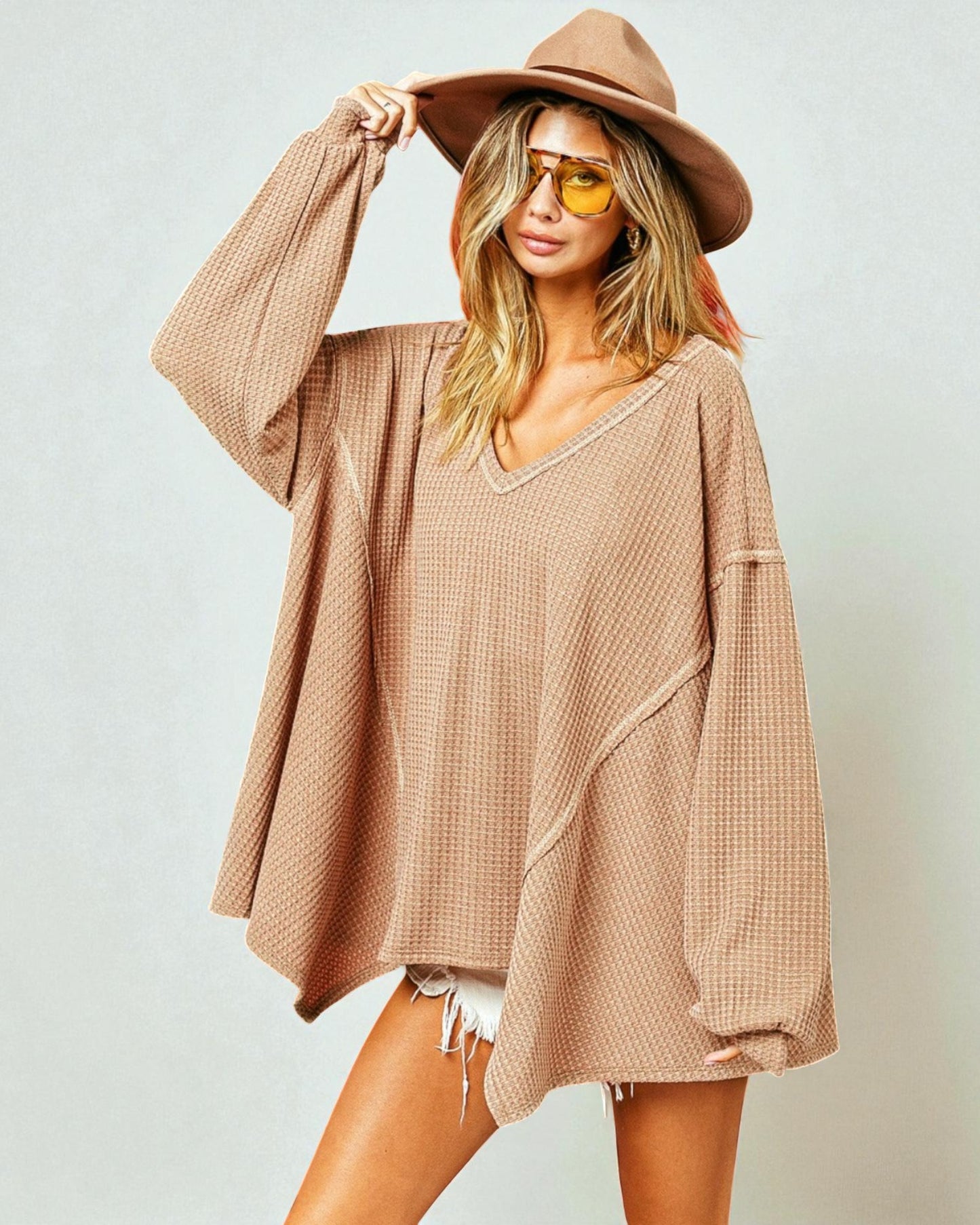 Woman wearing a beige oversized sweater and hat with sunglasses on a plain background