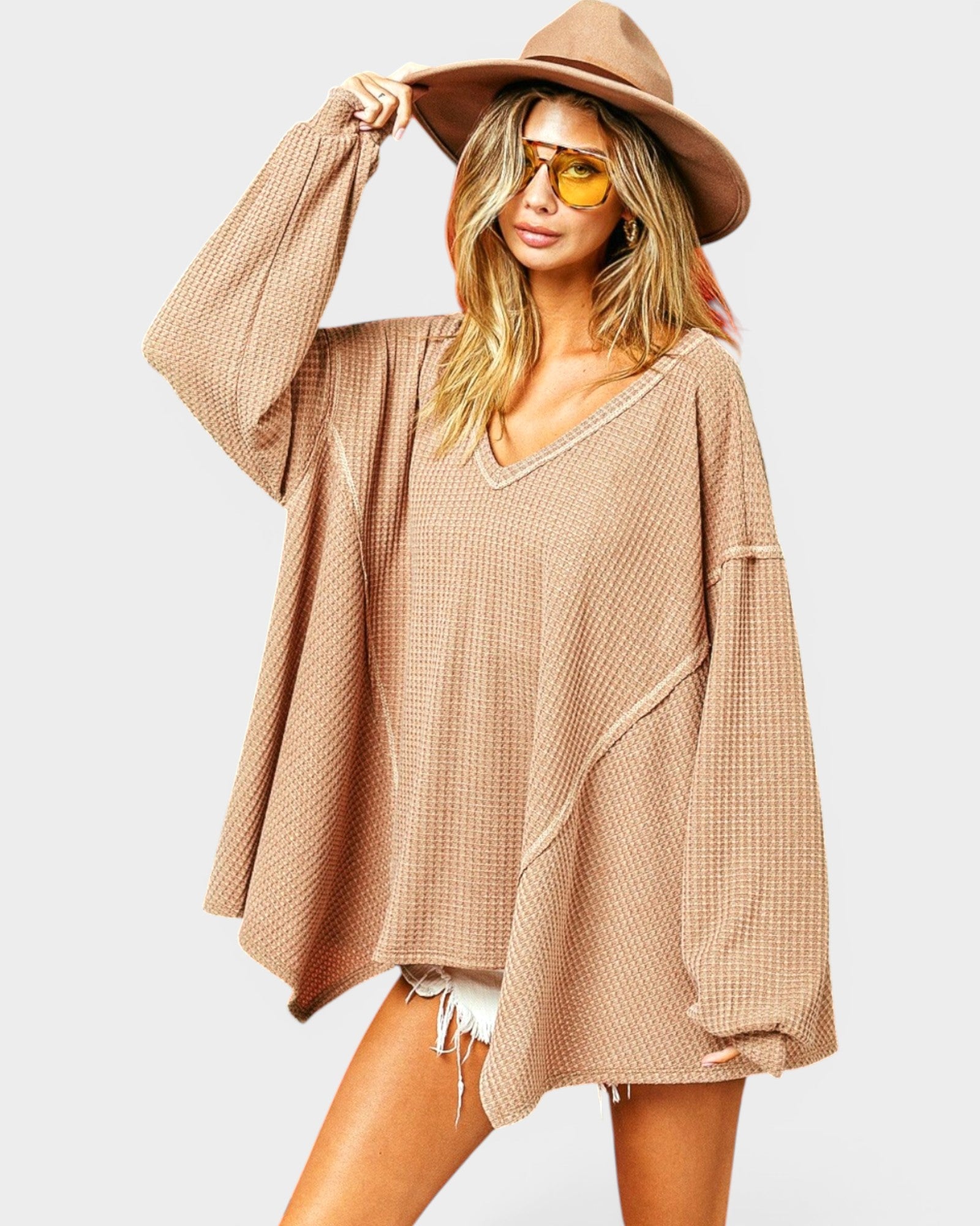 Woman wearing a beige oversized sweater, white shorts, and a wide-brimmed hat on a plain background