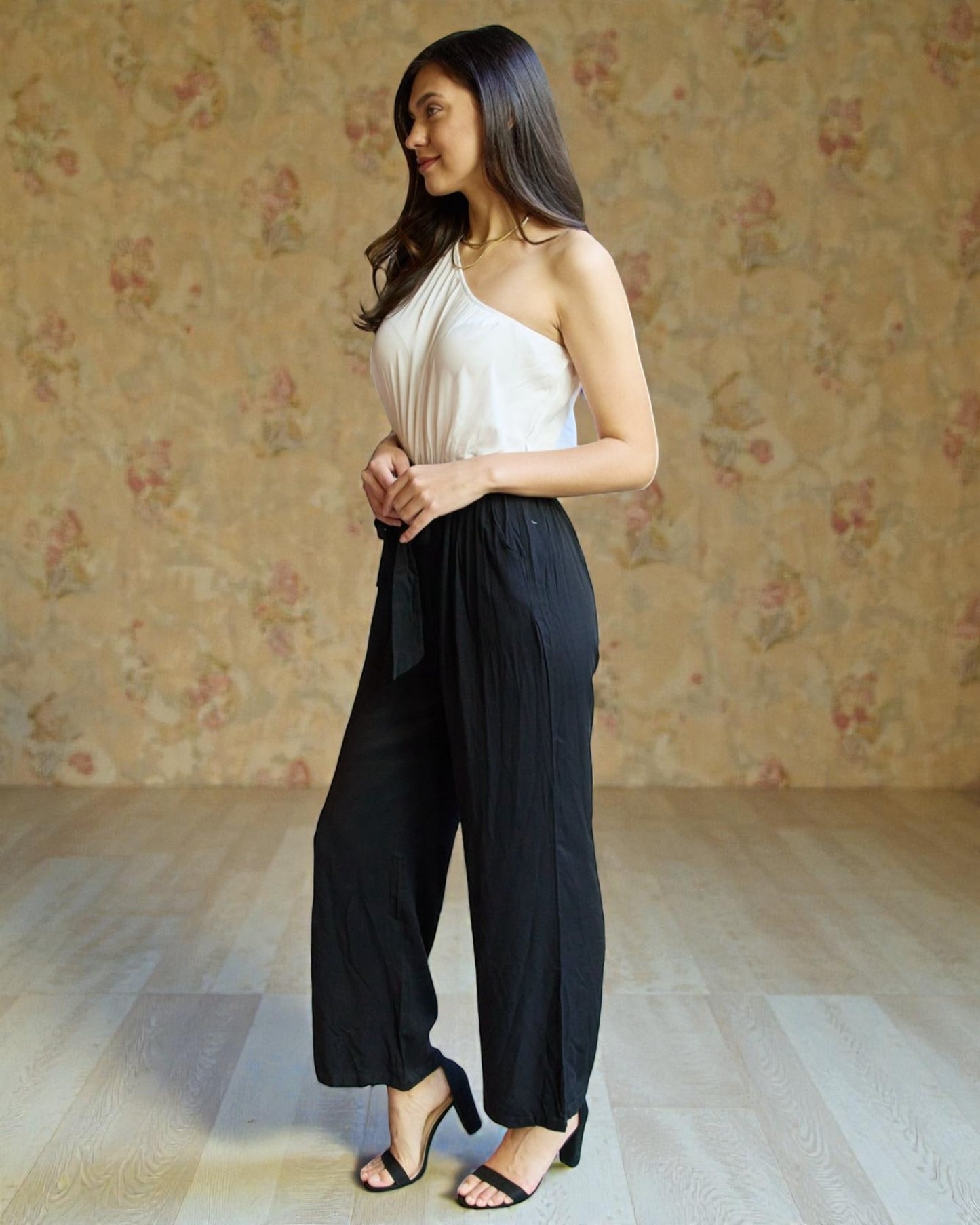 Woman wearing a white top and black pants standing against a floral wallpapered wall.