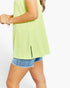 A woman wearing a sleeveless lime green ribbed tunic top with a side slit and denim shorts, shown from the side against a plain white background.