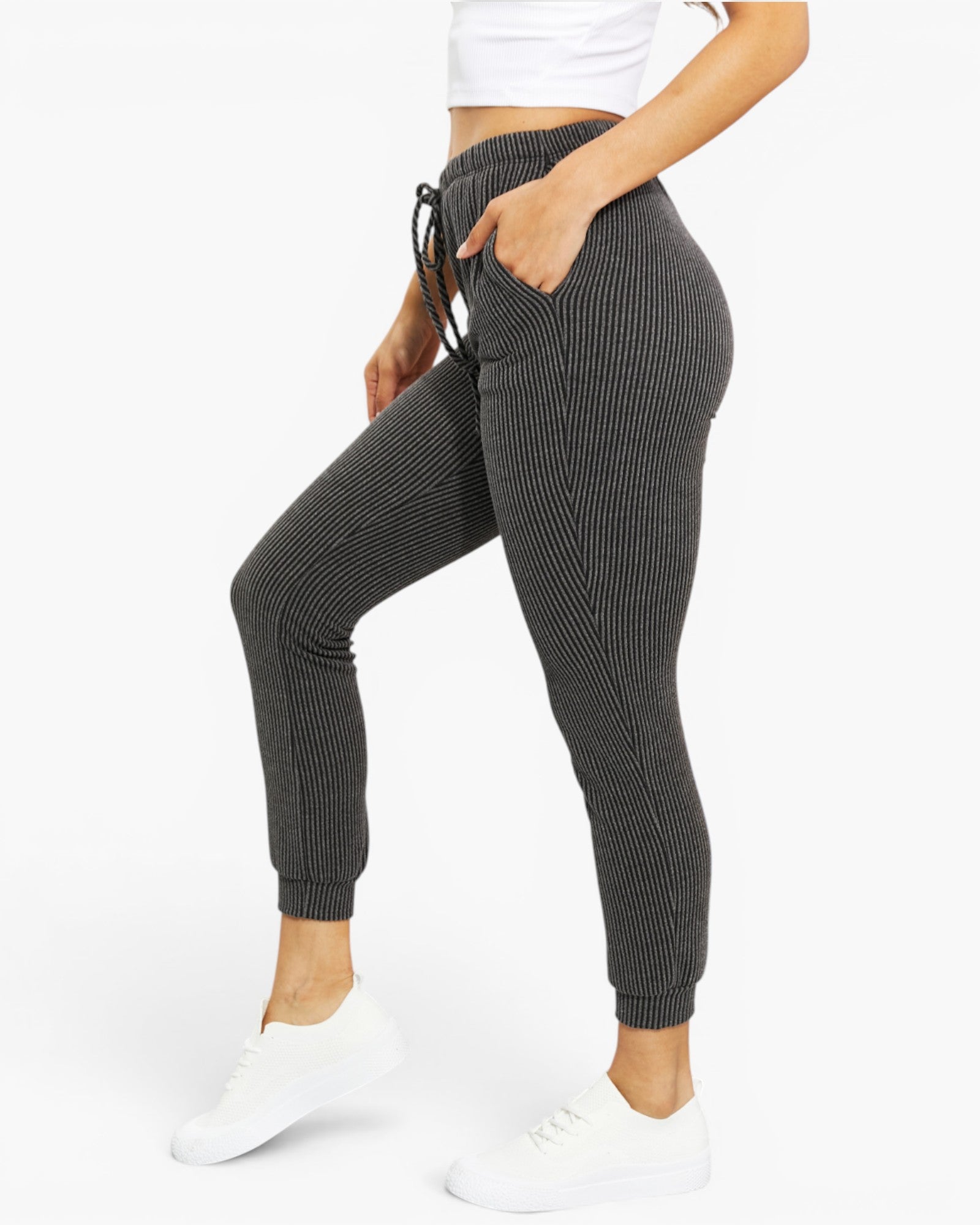 A model wearing dark gray ribbed jogger pants with a drawstring waist and side pockets, paired with a white crop top and white sneakers, shown from the waist down on a white studio background.