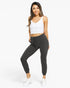 A woman modeling dark gray ribbed jogger pants with a drawstring waist, paired with a white crop top and white sneakers, against a light gray studio background.