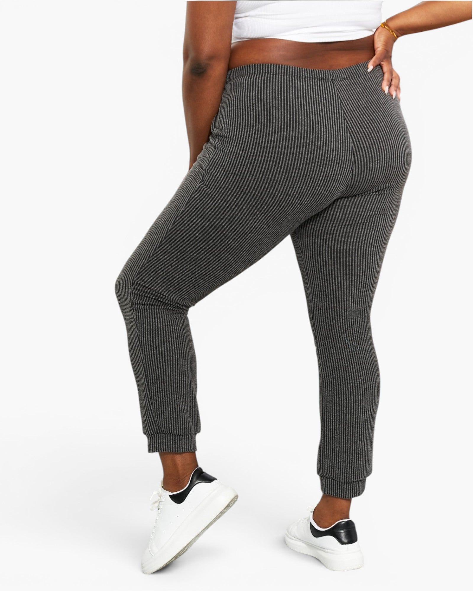 A person wearing dark gray ribbed jogger pants with a white crop top and white sneakers, shown from the waist down against a light gray studio background.