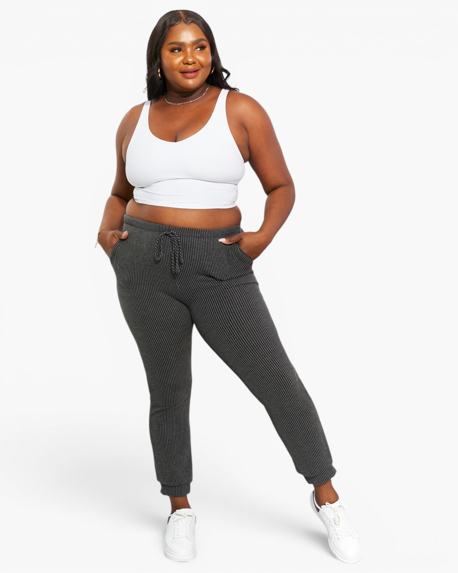 A plus-size model wearing dark gray ribbed jogger pants with a drawstring waist, paired with a white crop top and white sneakers, against a solid white studio background.