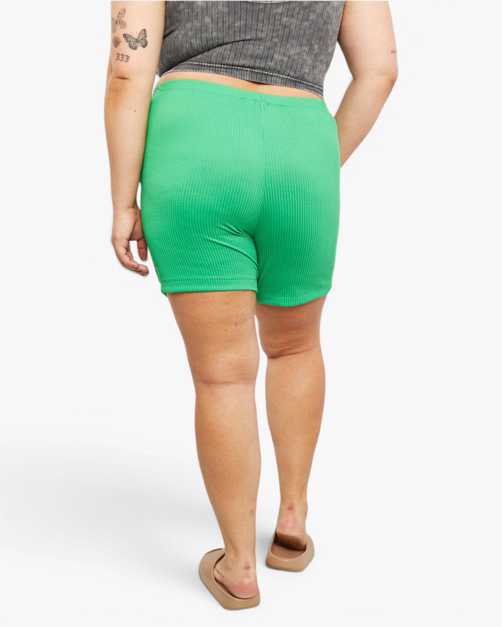 A person shown from the back wearing bright green ribbed shorts, a gray cropped top, and tan slide sandals against a plain light gray studio background.