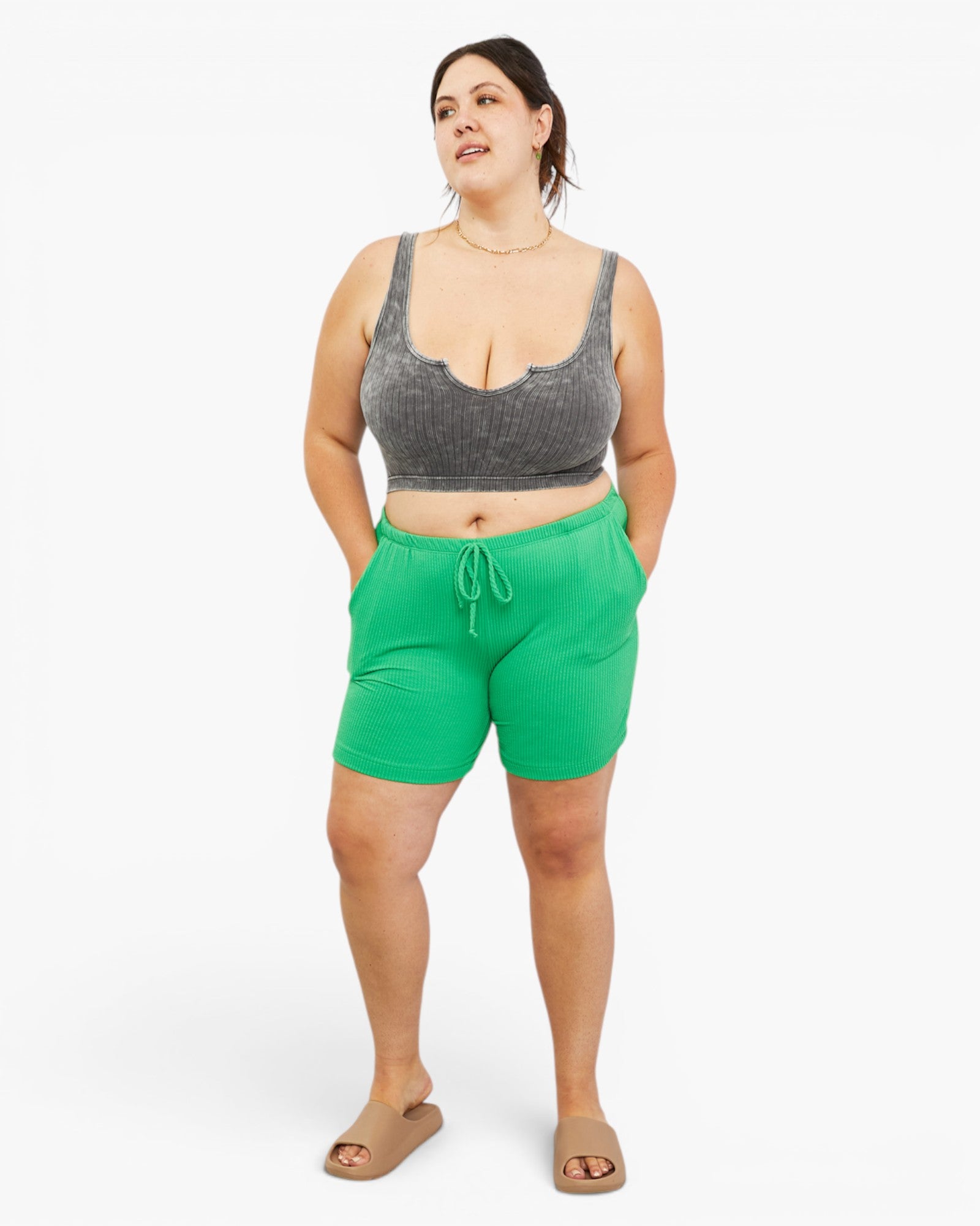 A woman modeling bright green drawstring shorts and a gray ribbed crop top against a plain light gray studio background.