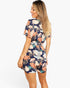 A woman modeling a short-sleeve floral romper from the back, featuring a navy blue base with orange, pink, and white tropical leaf patterns, against a plain white background.