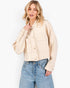 A woman wearing a cropped, light beige denim jacket with front buttons and flap pockets, paired with light blue jeans, against a plain white studio background.
