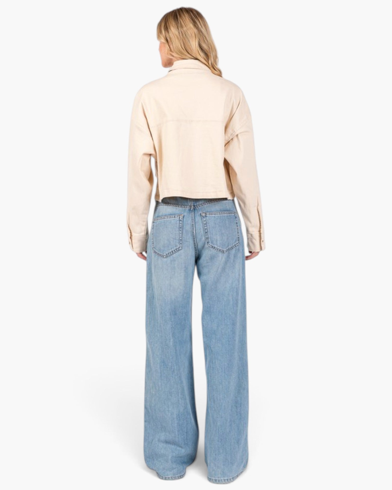 A woman shown from the back wearing a light beige cropped button-up shirt and light blue wide-leg jeans, photographed against a clean white studio background.