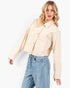A woman modeling a cropped, long-sleeve utility jacket in a light cream color, paired with light-wash blue jeans, against a plain white studio background.