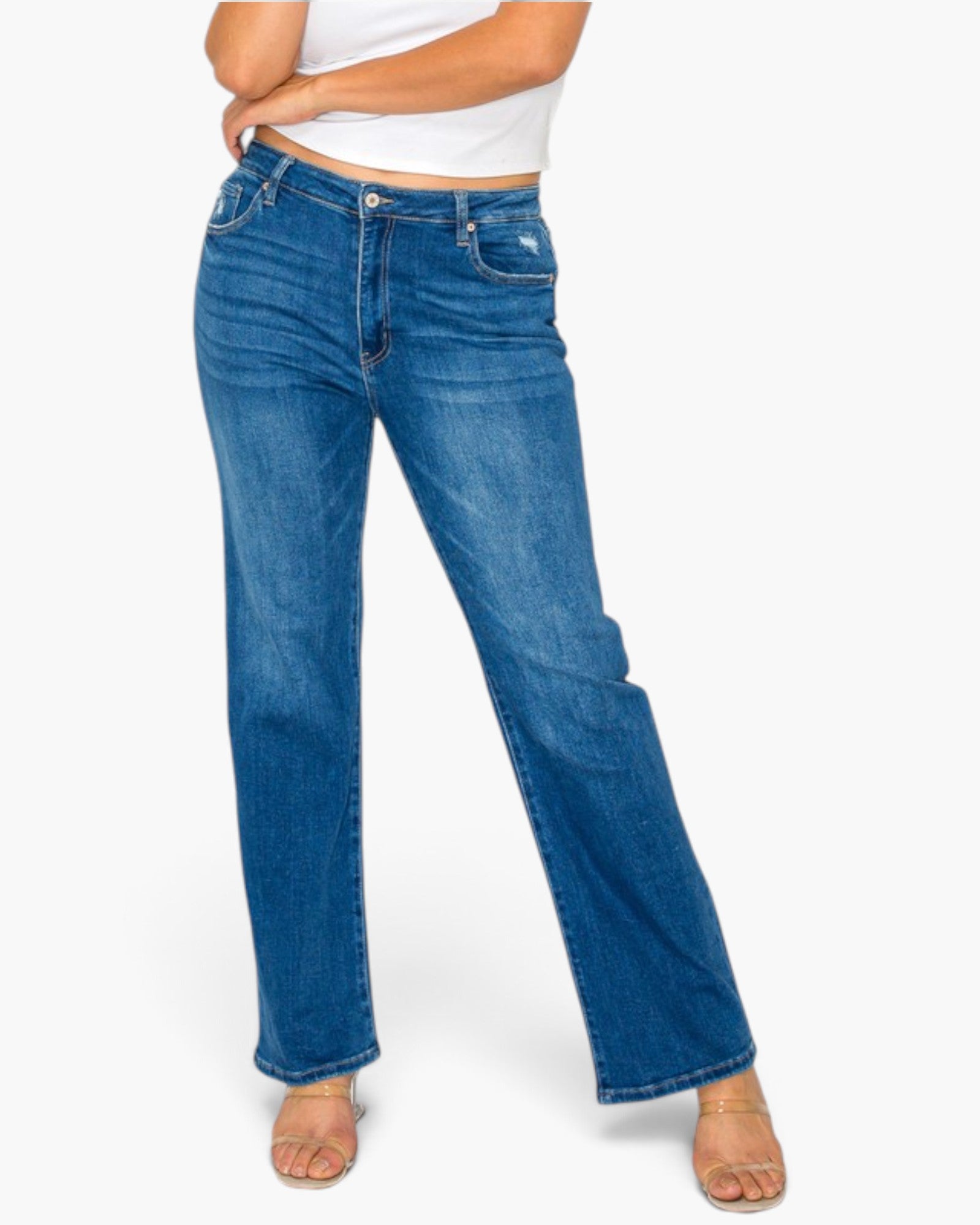 A model wearing a pair of medium wash blue denim jeans with a straight-to-medium leg fit, styled with a white crop top and clear high-heeled sandals against a light gray background.