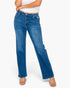 A model wearing a pair of medium wash blue denim jeans with a straight-to-medium leg fit, styled with a white crop top and clear high-heeled sandals against a light gray background.