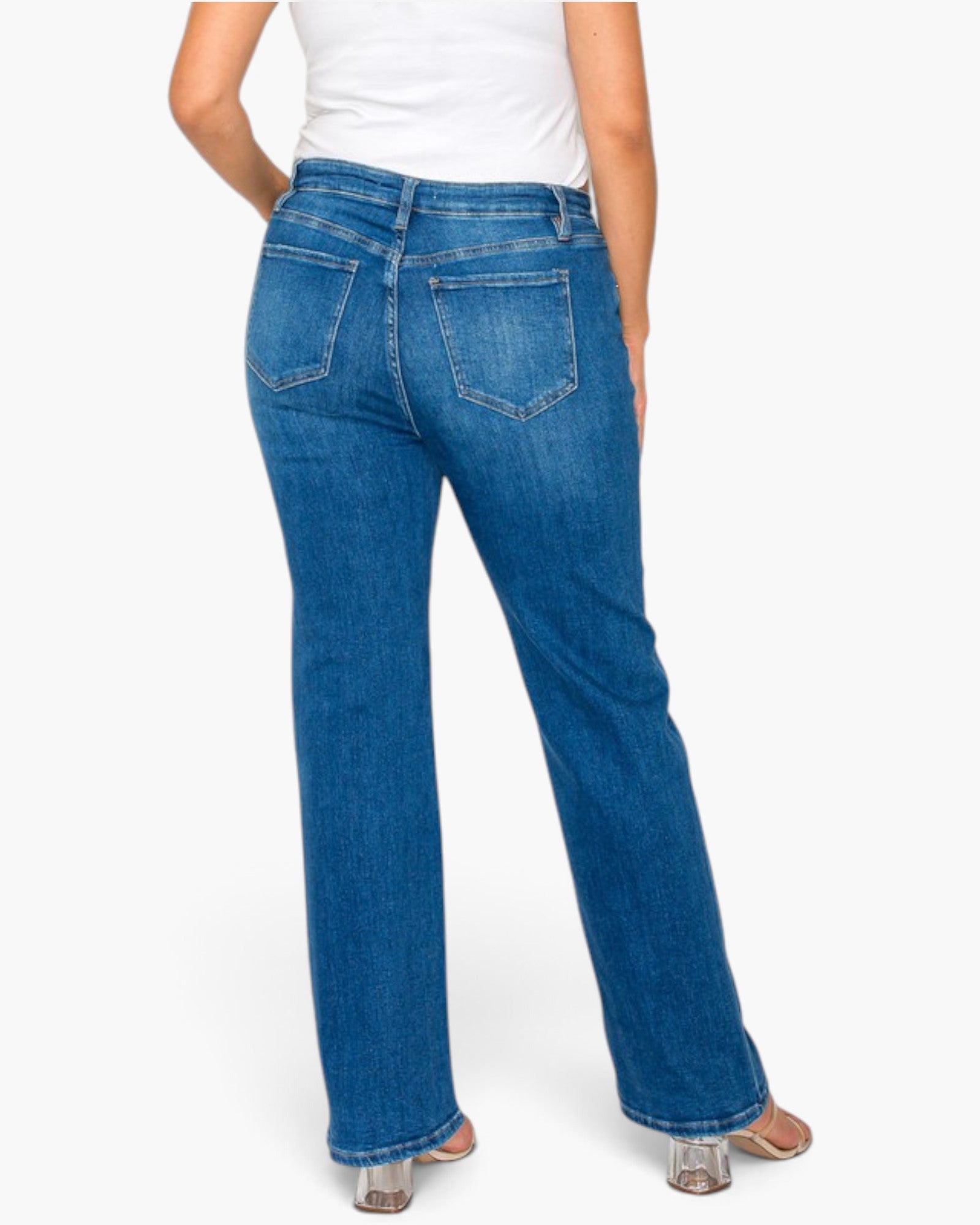 Rear view of a person wearing blue denim jeans with a straight-leg fit, paired with a white top and clear high-heeled sandals, against a light gray studio background.