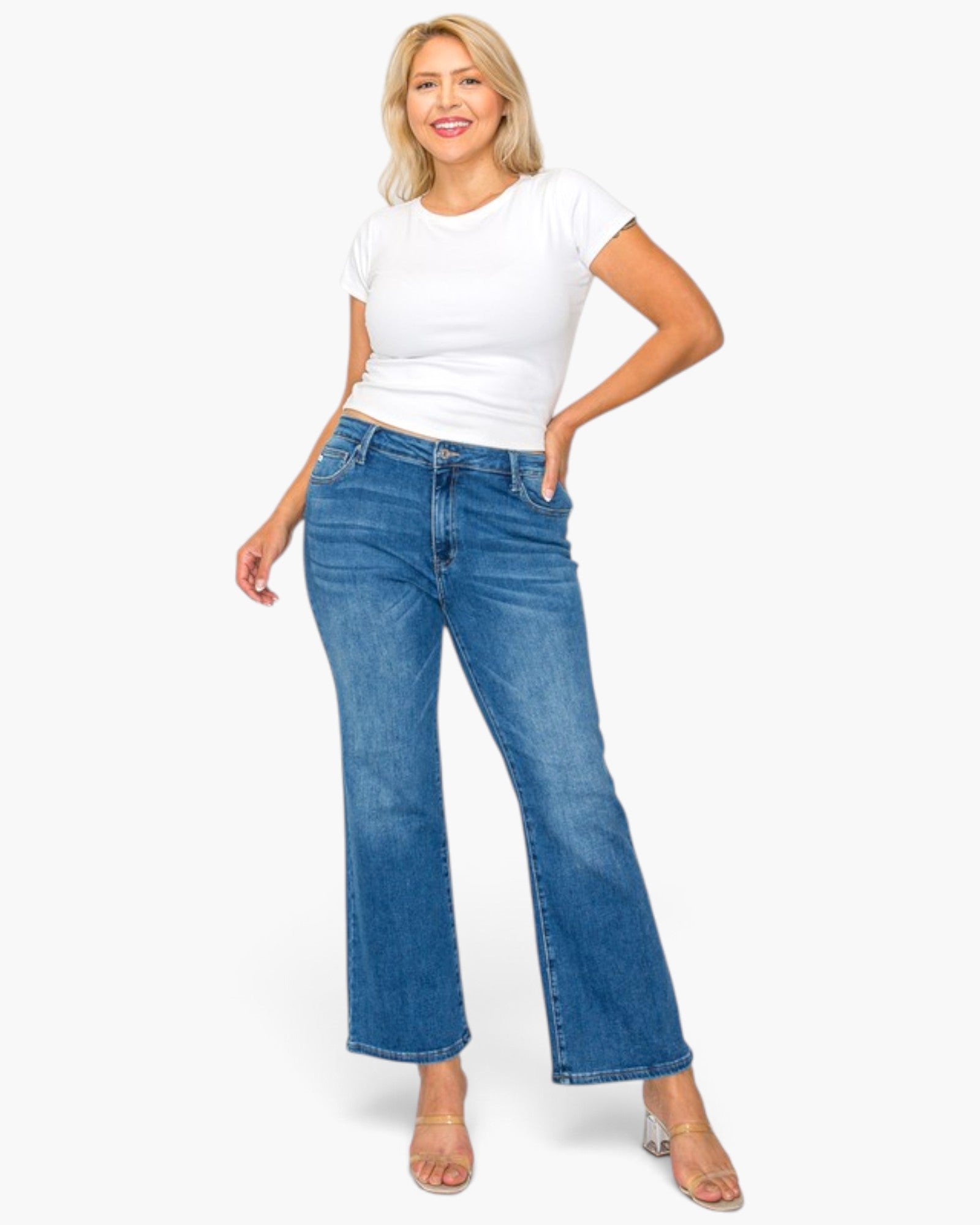 A woman modeling a pair of medium-wash blue flare jeans with a white t-shirt and clear high-heeled sandals, standing against a solid white background.