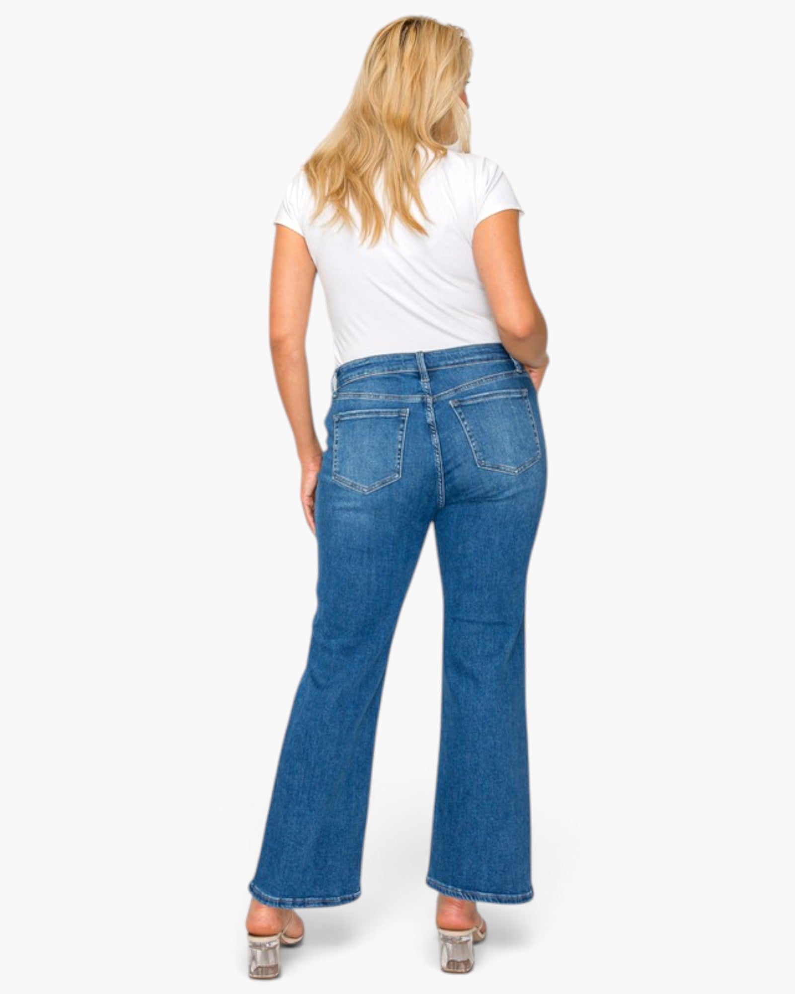 A woman viewed from behind wearing blue flare jeans, a white t-shirt, and metallic high heels against a plain white background.