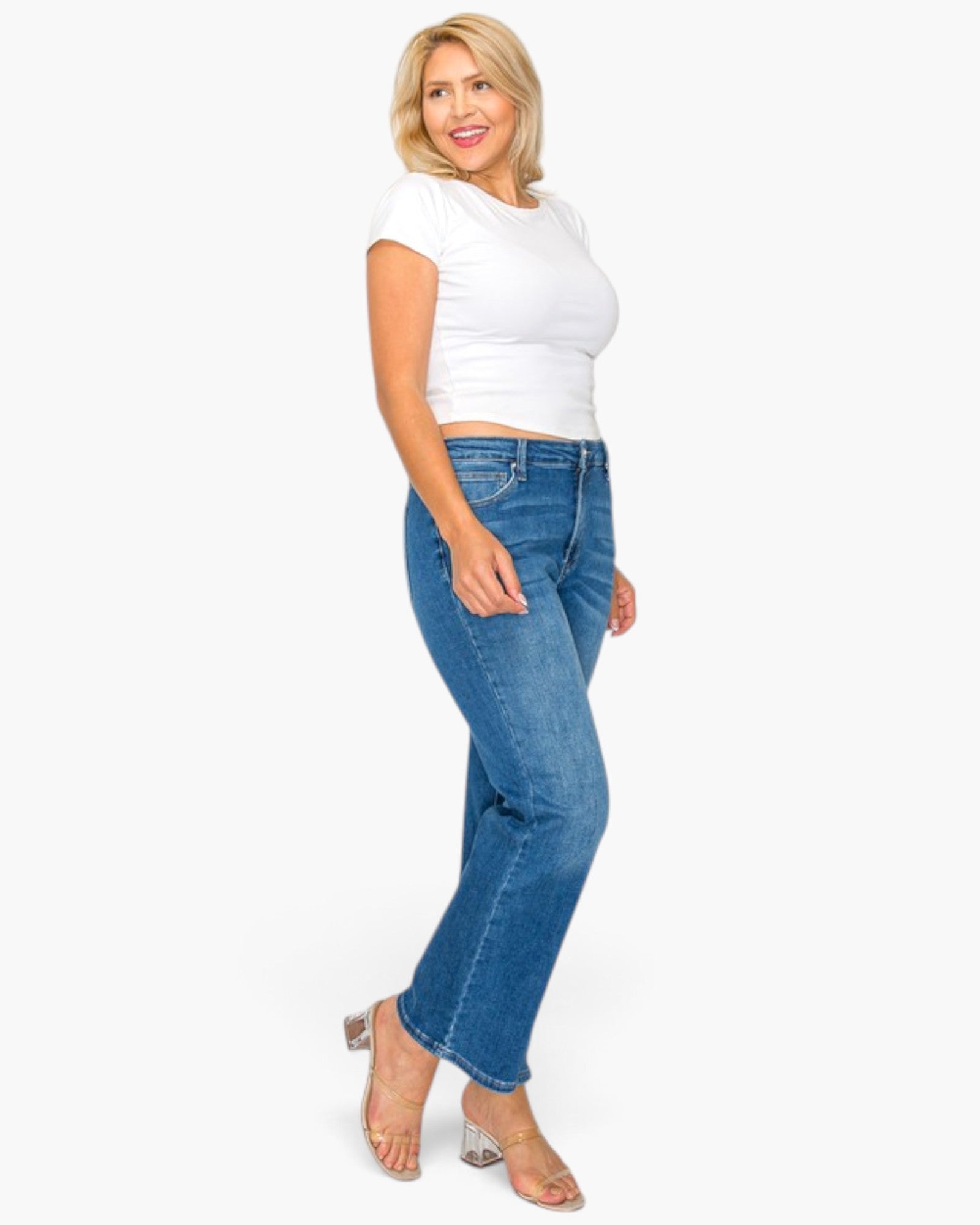A woman modeling a pair of medium-wash blue jeans with a white cropped t-shirt and clear high-heeled sandals, standing against a solid white background.