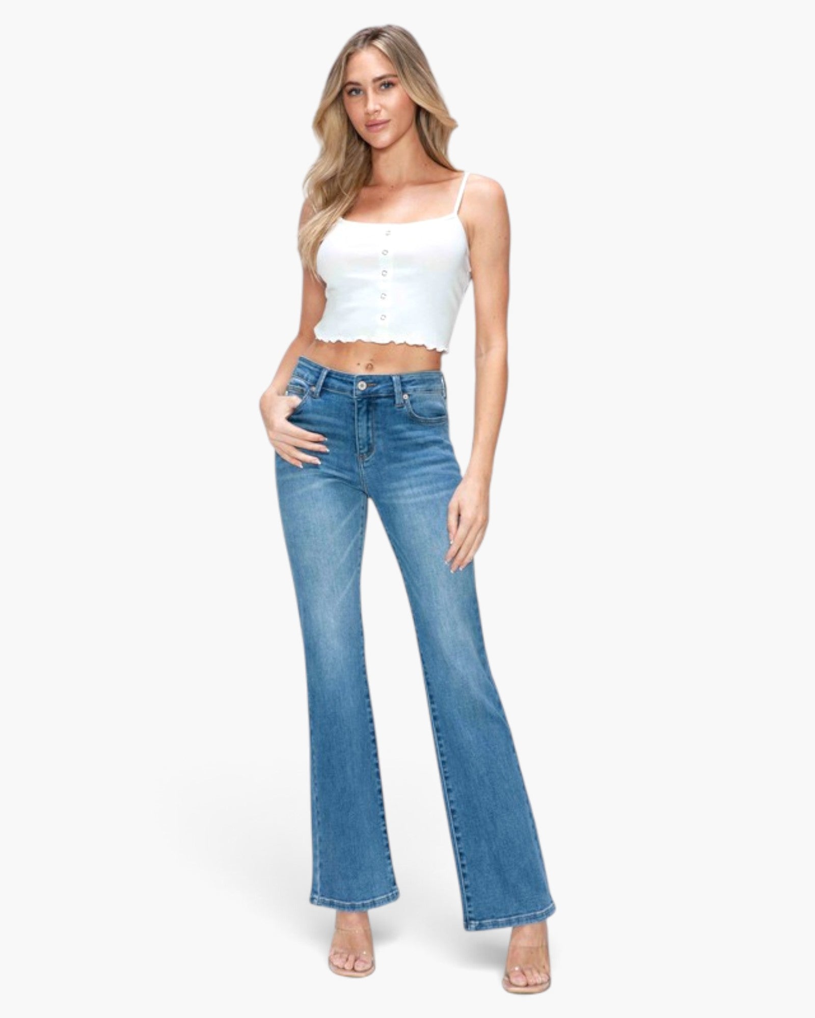 A woman modeling a pair of high-waisted, blue flare jeans with a white button-up crop top against a plain white studio background.