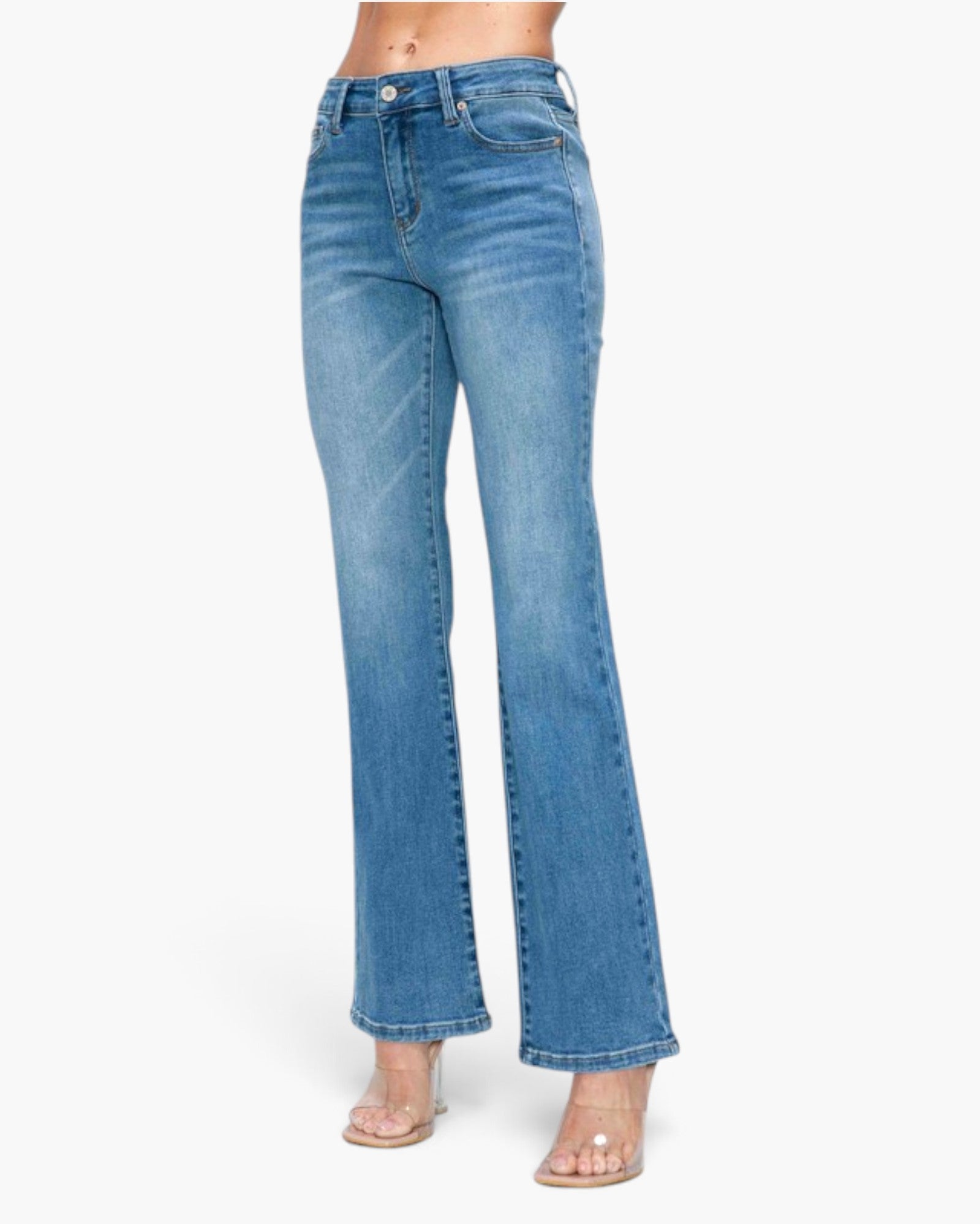 A model from the waist down wearing a pair of medium-wash blue denim flare jeans with clear high-heeled sandals, set against a plain white studio background.