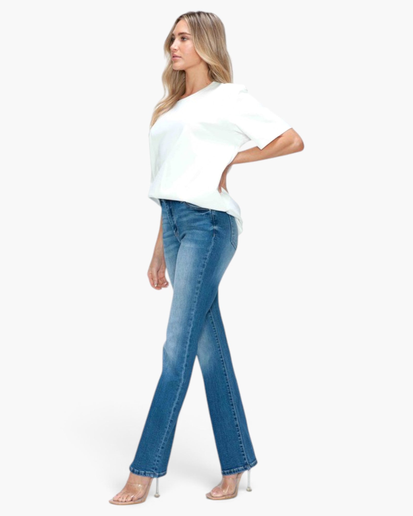 A woman modeling blue flare jeans with a white t-shirt and clear high-heeled sandals, shown from a side profile against a plain white background.