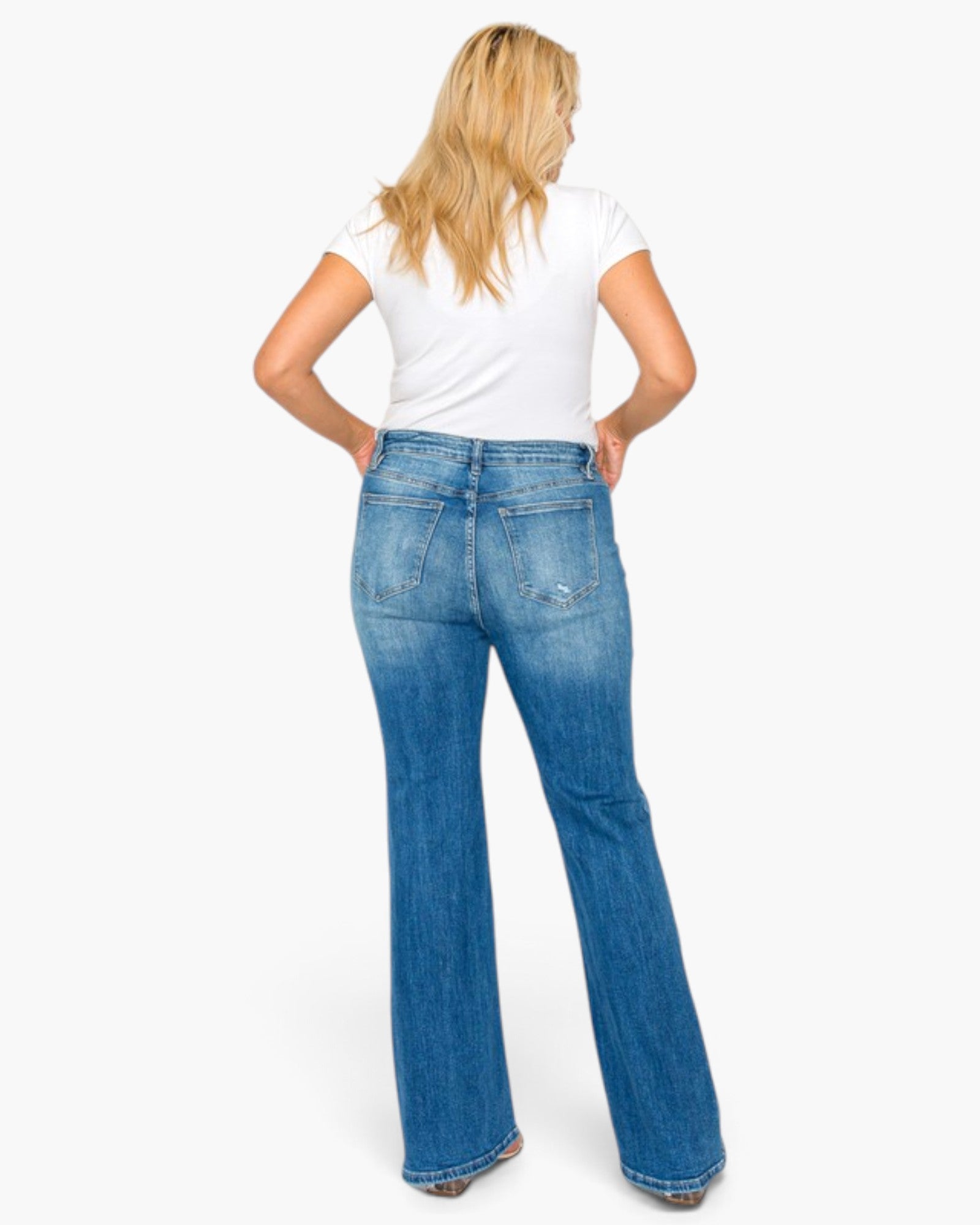A woman viewed from behind wearing blue flare jeans and a white t-shirt, standing against a plain white studio background.
