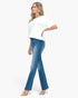 A woman modeling blue flare jeans with a white t-shirt and clear high-heeled sandals, shown from a side profile against a plain white background.
