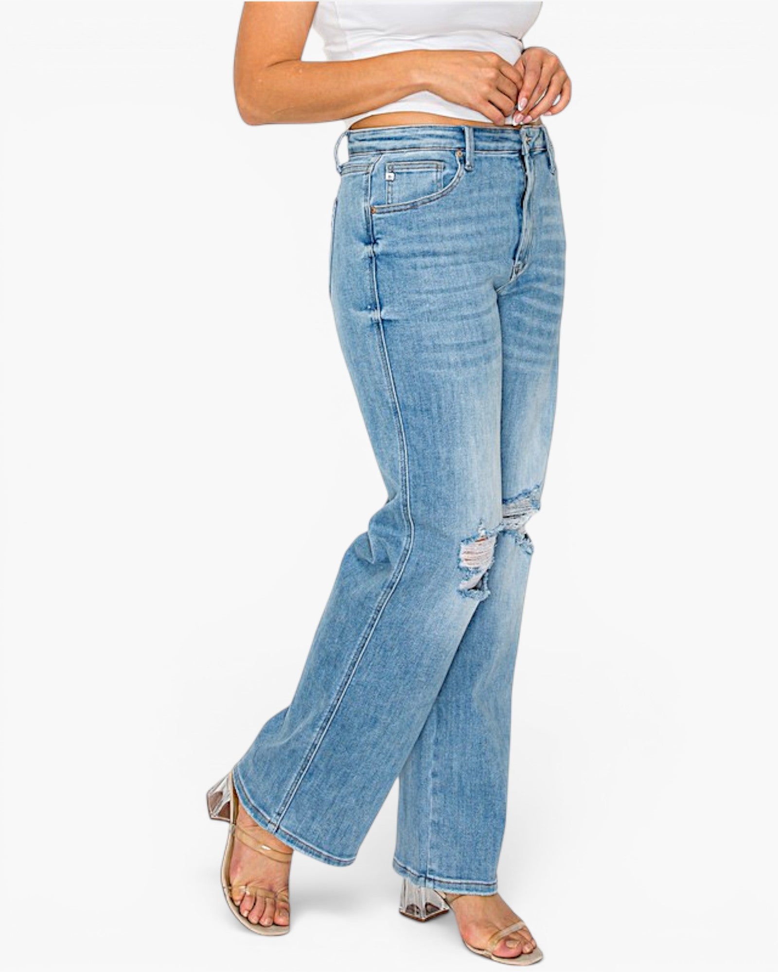 A model wearing light-wash blue distressed jeans with a wide leg and ripped details on the knees, paired with a white crop top and metallic sandals, against a plain white background.