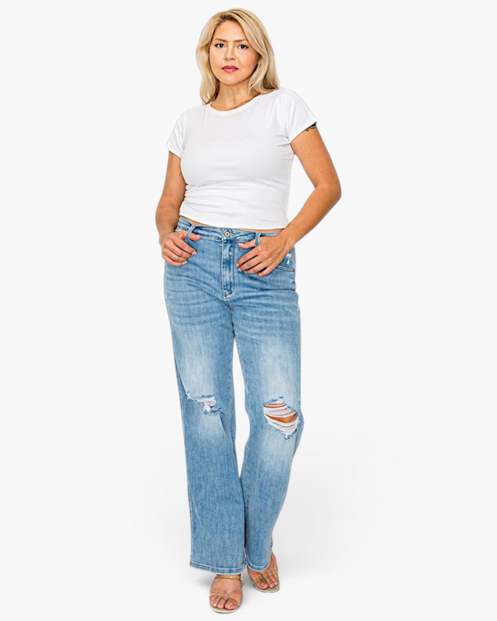A woman modeling a pair of light-wash blue distressed jeans with a white t-shirt, standing against a plain white studio background.