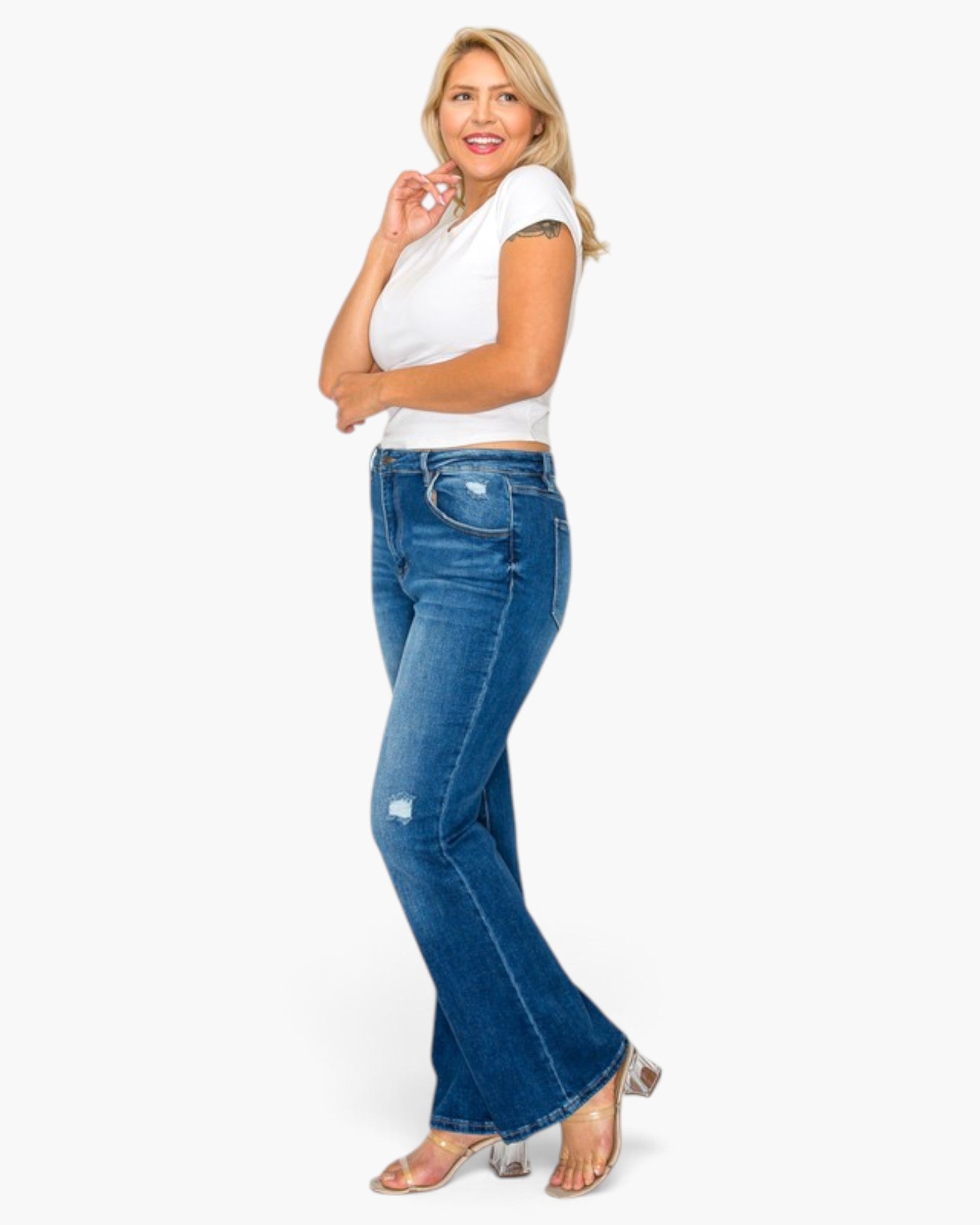 A woman modeling a pair of blue high-waisted flare jeans with a white crop top and clear high-heeled sandals, standing against a plain white background.