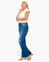 A woman modeling a pair of blue high-waisted flare jeans with a white crop top and clear high-heeled sandals, standing against a plain white background.