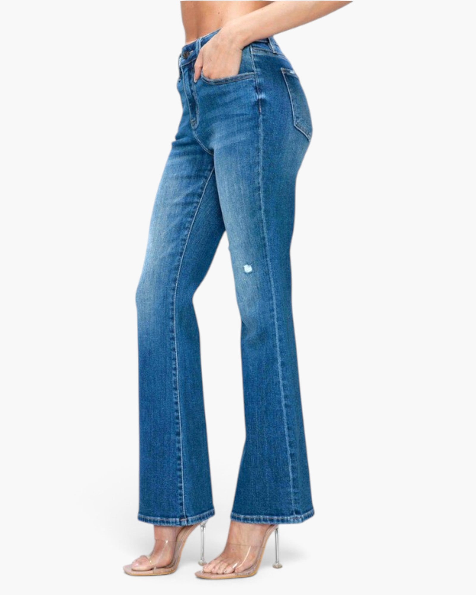 A model wearing blue high-waisted flare jeans with a small distressed detail on the upper leg, paired with clear high-heeled sandals, shown against a plain white background.