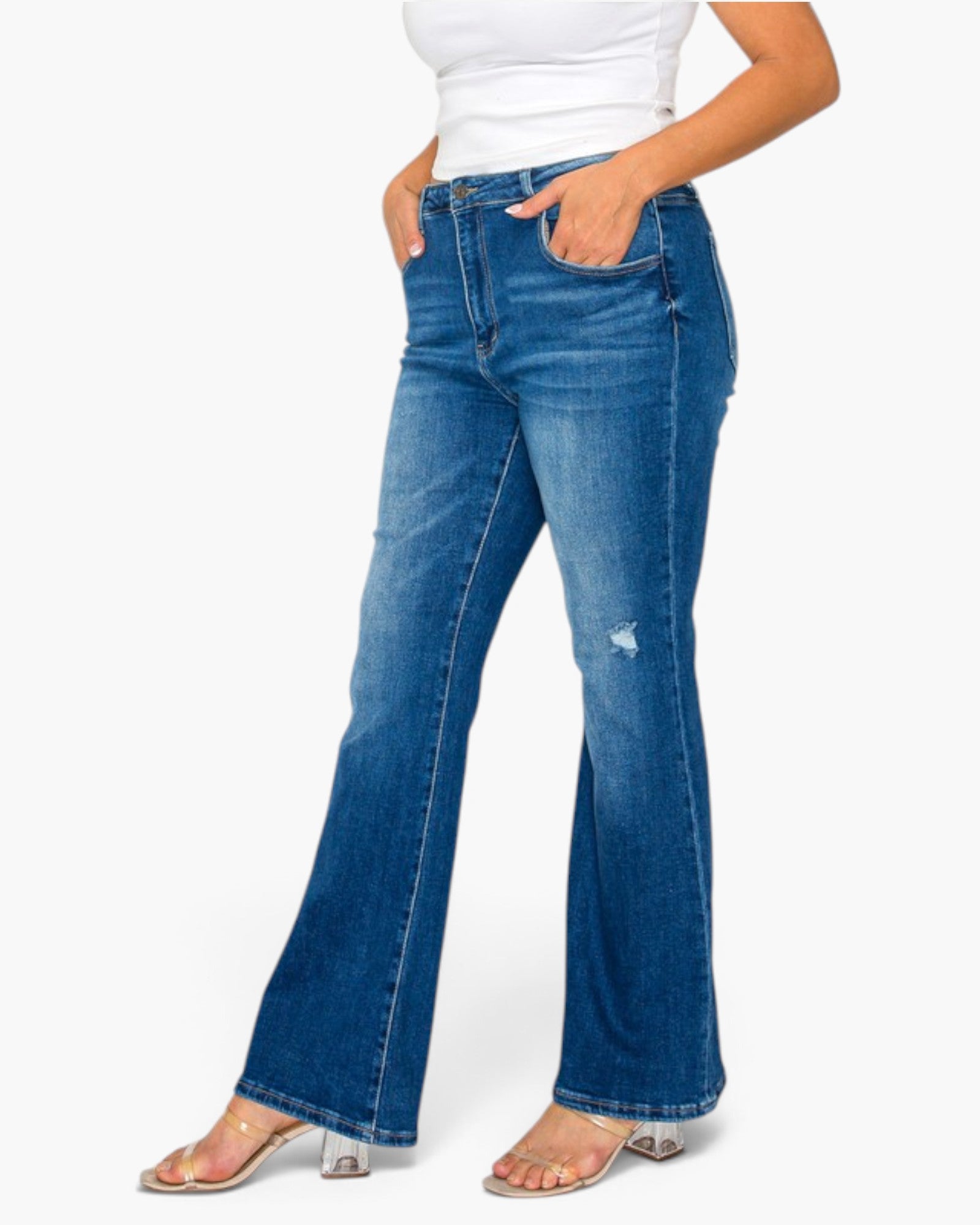 A person wearing medium-wash blue flare jeans with a small distressed detail on one leg, paired with a white top and clear high-heeled sandals, against a plain white background.