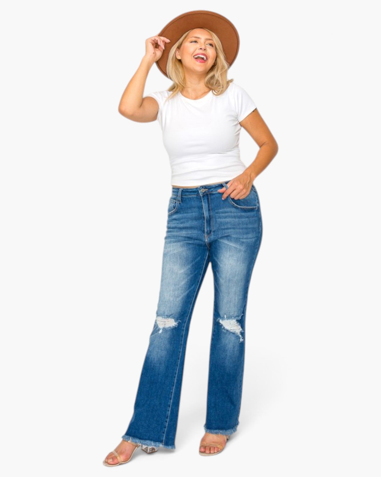 A woman modeling a pair of blue distressed flare jeans with a white t-shirt and a brown wide-brim hat, standing against a light gray background.