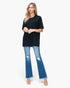 A woman modeling a black short-sleeve tunic top paired with blue distressed flare jeans and clear high-heeled sandals, photographed against a plain light gray studio background.
